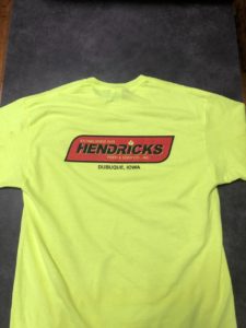 Photo of Hendricks Feed T-shirt. The shirt is yellow with the red Hendricks logo across the front. Written in black and yellow on the red is '(yellow) Estabished 1929, (black) Hendricks, (yellow) Feed & Seed Co., Inc.' is in the banner and 'Dubuque, Iowa' is below the banner.